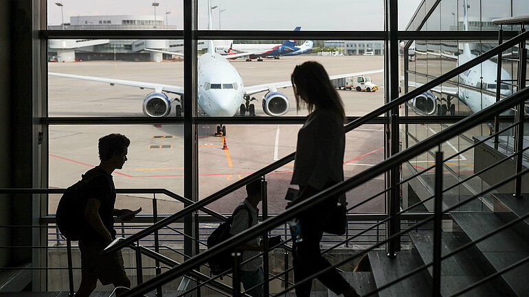 Two passengers in the foreground are walking up the stairs of an airport gate. A passenger aircraft can be seen through a large window. Two passengers in the foreground.