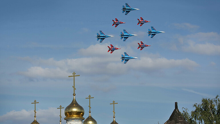 Nine blue and red fighter jets fly in rectangular formation over the golden onion domes of an Orthodox church.